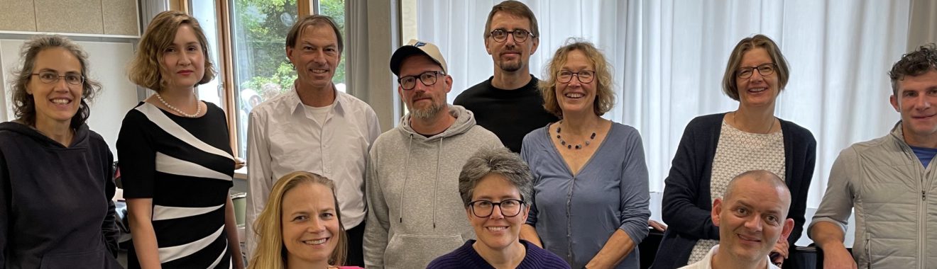 Eine Gruppe von zwölf Personen steht und sitzt in einem hellen Raum mit Fenstern und Vorhängen. Die Personen sind unterschiedlich gekleidet und lächeln in die Kamera. Im Vordergrund sitzen drei Personen, während die anderen im Hintergrund stehen. Der Raum hat einen neutralen Boden und eine schlichte Einrichtung.