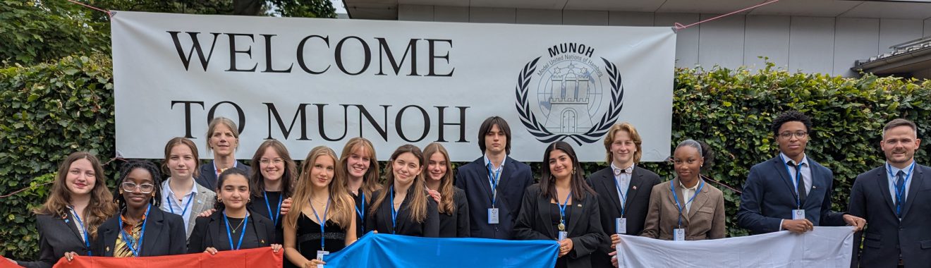 Eine Gruppe von Jugendlichen steht vor einem Banner mit der Aufschrift "WELCOME TO MUNOH". Sie halten Flaggen in den Farben Blau, Weiß und Rot. Die Personen tragen formelle Kleidung und lächeln in die Kamera. Im Hintergrund ist eine grüne Hecke zu sehen.