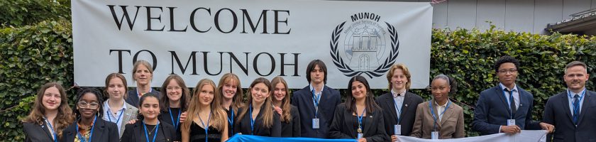 Eine Gruppe von Jugendlichen steht vor einem Banner mit der Aufschrift "WELCOME TO MUNOH". Sie halten Flaggen in den Farben Blau, Weiß und Rot. Die Personen tragen formelle Kleidung und lächeln in die Kamera. Im Hintergrund ist eine grüne Hecke zu sehen.
