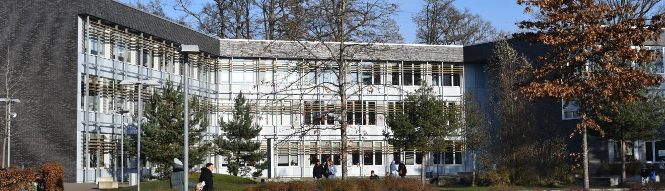 Moderne Gebäudefront mit großen Fenstern und Glasfassade, umgeben von Bäumen und Sträuchern. Im Vordergrund sind einige Menschen zu sehen, die sich im Freien aufhalten. Der Himmel ist blau und sonnig.