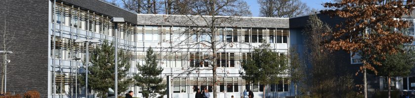Moderne Gebäudefront mit großen Fenstern und Glasfassade, umgeben von Bäumen und Sträuchern. Im Vordergrund sind einige Menschen zu sehen, die sich im Freien aufhalten. Der Himmel ist blau und sonnig.