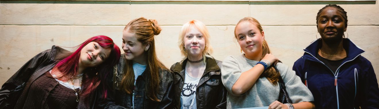 Fünf junge Frauen posieren vor einem Schild mit der Aufschrift "DIE ZEIT". Sie halten ein Banner mit dem Hashtag "#ICHSTEHauf". Die Umgebung wirkt modern und einladend. Die Frauen zeigen verschiedene Posen und Gesichtsausdrücke, die Freude und Zusammenhalt ausstrahlen.