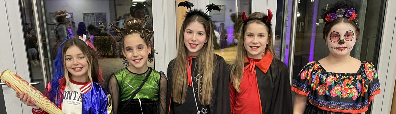 Fünf Mädchen in bunten Kostümen stehen nebeneinander vor einem Fenster. Sie lächeln und posieren. Jedes Mädchen trägt ein einzigartiges Outfit, das Elemente von Halloween und traditionellen Feierlichkeiten kombiniert. Im Hintergrund sind unscharfe Lichter zu sehen.