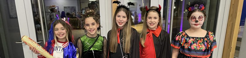 Fünf Mädchen in bunten Kostümen stehen nebeneinander vor einem Fenster. Sie lächeln und posieren. Jedes Mädchen trägt ein einzigartiges Outfit, das Elemente von Halloween und traditionellen Feierlichkeiten kombiniert. Im Hintergrund sind unscharfe Lichter zu sehen.