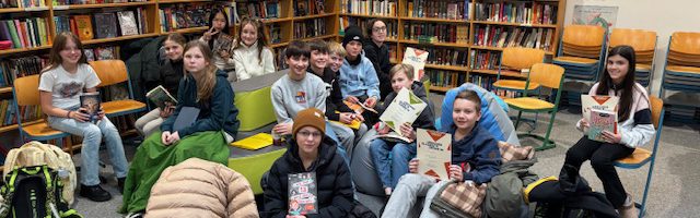 Eine Gruppe von Jugendlichen sitzt in einer Bibliothek auf Stühlen und Sofas. Sie halten Bücher in den Händen und schauen in die Kamera. Im Hintergrund sind Regale mit vielen Büchern zu sehen. Die Atmosphäre ist entspannt und freundlich.