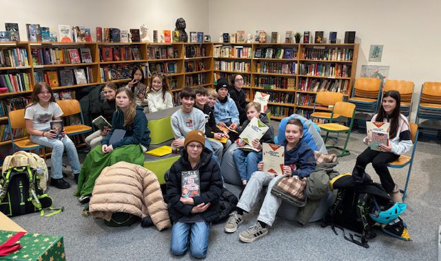 Eine Gruppe von Jugendlichen sitzt in einer Bibliothek auf Stühlen und Sofas. Sie halten Bücher in den Händen und schauen in die Kamera. Im Hintergrund sind Regale mit vielen Büchern zu sehen. Die Atmosphäre ist entspannt und freundlich.