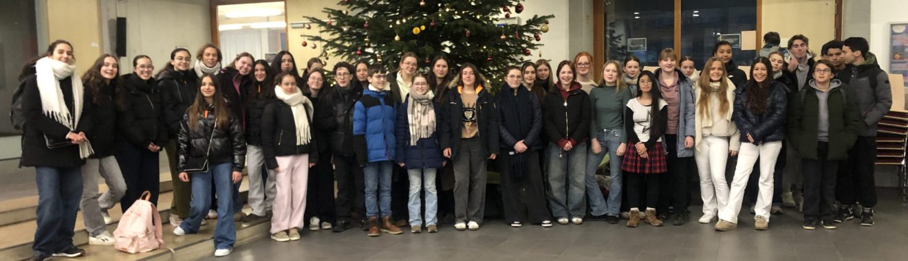 Gruppenfoto von etwa 50 Personen, die in einem Innenraum vor einem Weihnachtsbaum stehen. Die Menschen sind unterschiedlich gekleidet und lächeln in die Kamera. Im Hintergrund sind Fenster und eine Wand zu sehen.