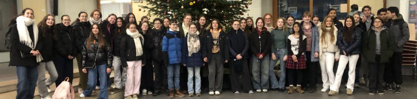 Gruppenfoto von etwa 50 Personen, die in einem Innenraum vor einem Weihnachtsbaum stehen. Die Menschen sind unterschiedlich gekleidet und lächeln in die Kamera. Im Hintergrund sind Fenster und eine Wand zu sehen.