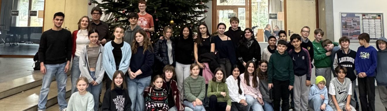 Gruppenfoto von Schülerinnen und Schülern in einer Halle, umgeben von großen Fenstern und einem Weihnachtsbaum. Die Personen stehen in mehreren Reihen und lächeln in die Kamera.