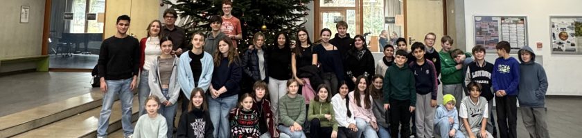 Gruppenfoto von Schülerinnen und Schülern in einer Halle, umgeben von großen Fenstern und einem Weihnachtsbaum. Die Personen stehen in mehreren Reihen und lächeln in die Kamera.