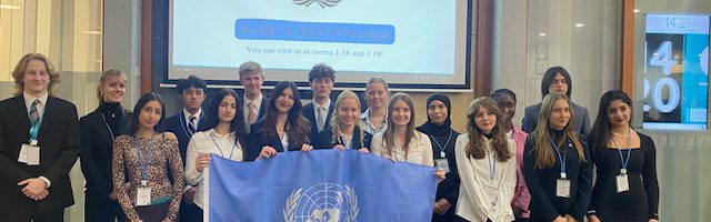 Gruppe von 15 Personen in formeller Kleidung, die zusammen vor einem Bildschirm mit dem Logo einer Organisation stehen. Eine Person hält eine Flagge der Vereinten Nationen. Im Hintergrund sind mehrere nationale Flaggen und ein Bildschirm mit Informationen zu sehen.