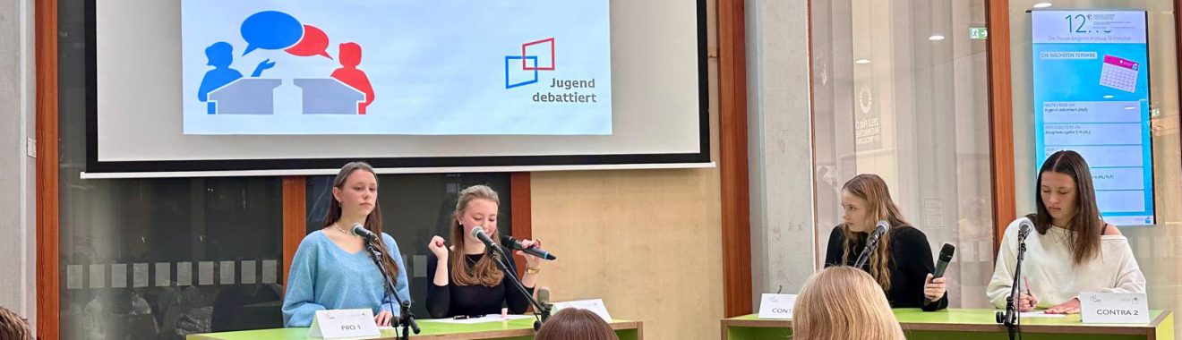 Eine Gruppe von vier Personen sitzt an Tischen, während sie an einer Diskussionsveranstaltung teilnehmen. Im Hintergrund ist eine große Präsentationsleinwand zu sehen, auf der der Titel "Schulfinale" steht. Die Anwesenden scheinen aufmerksam zuzuhören. Einige Teilnehmer sitzen im Publikum und blicken in Richtung der Sprecher.