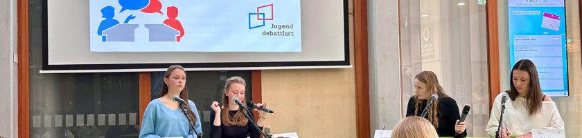 Eine Gruppe von vier Personen sitzt an Tischen, während sie an einer Diskussionsveranstaltung teilnehmen. Im Hintergrund ist eine große Präsentationsleinwand zu sehen, auf der der Titel "Schulfinale" steht. Die Anwesenden scheinen aufmerksam zuzuhören. Einige Teilnehmer sitzen im Publikum und blicken in Richtung der Sprecher.