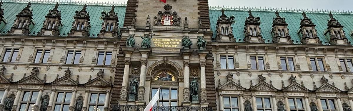 Gruppierung von Menschen vor dem Hamburger Rathaus, das im Hintergrund zu sehen ist. Das Gebäude hat eine beeindruckende Architektur mit einem hohen Turm und einer Flagge, die weht. Der Himmel ist bewölkt und es scheint, als wäre es kühl.