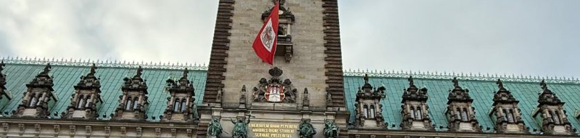 Gruppierung von Menschen vor dem Hamburger Rathaus, das im Hintergrund zu sehen ist. Das Gebäude hat eine beeindruckende Architektur mit einem hohen Turm und einer Flagge, die weht. Der Himmel ist bewölkt und es scheint, als wäre es kühl.