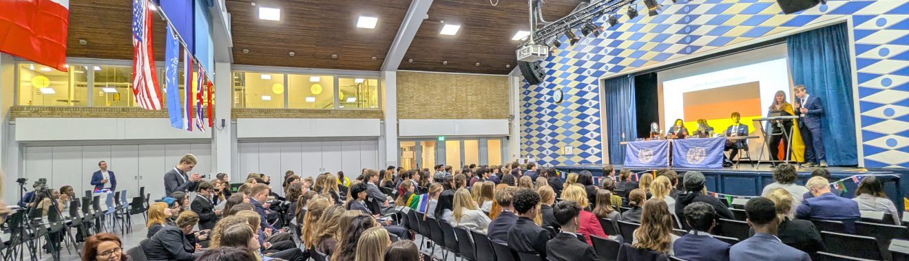 Eine große Gruppe von Menschen sitzt in einem Auditorium mit vielen leeren Stühlen. Auf der Bühne sind Redner zu sehen, während im Hintergrund verschiedene Flaggen hängen. Die Raumdecke ist hoch und die Wände sind mit einem geometrischen Muster dekoriert. Das Licht ist hell und es scheint eine Veranstaltung oder Konferenz stattzufinden.