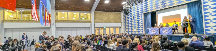 Eine große Gruppe von Menschen sitzt in einem Auditorium mit vielen leeren Stühlen. Auf der Bühne sind Redner zu sehen, während im Hintergrund verschiedene Flaggen hängen. Die Raumdecke ist hoch und die Wände sind mit einem geometrischen Muster dekoriert. Das Licht ist hell und es scheint eine Veranstaltung oder Konferenz stattzufinden.