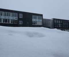 Schneebedeckte Fläche im Vordergrund mit zwei modernen Gebäuden im Hintergrund, unter einem grauen, bewölkten Himmel.