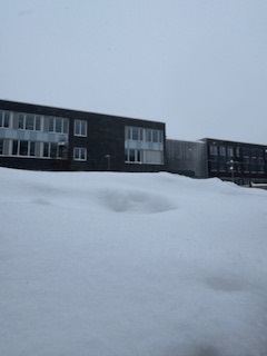 Schneebedeckte Fläche im Vordergrund mit zwei modernen Gebäuden im Hintergrund, unter einem grauen, bewölkten Himmel.
