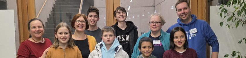 Eine Gruppe von neun Personen steht in einem Innenraum. Sie lächeln in die Kamera. Die Gruppe setzt sich aus Erwachsenen und Kindern unterschiedlicher Altersgruppen zusammen. Im Hintergrund sind Fenster und Pflanzen zu sehen. Einige Personen tragen Namensschilder.