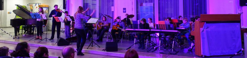 Eine Gruppe von Musikern spielt auf einer Bühne. Im Vordergrund ist eine Musikerin mit einem Instrument zu sehen, während im Hintergrund weitere Musiker an verschiedenen Instrumenten, darunter ein Klavier und eine Gitarre, sitzen. Die Beleuchtung ist in lila Tönen gehalten und schafft eine festliche Atmosphäre. Zuschauer sind im unteren Bildbereich sichtbar.