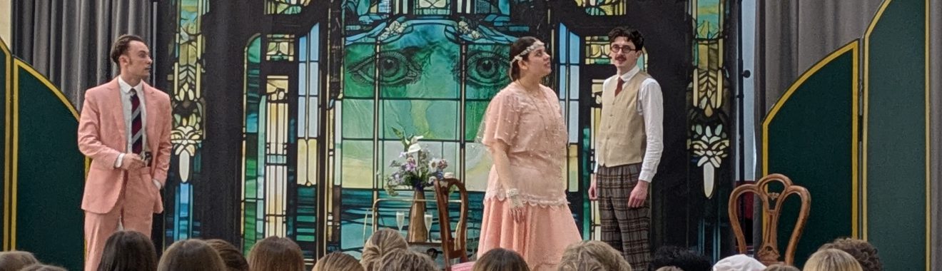 Drei Schauspieler stehen auf einer Bühne, während das Publikum im Vordergrund sitzt. Die beiden männlichen Darsteller tragen Anzüge in Pastellfarben, während die weibliche Darstellerin ein elegantes, helles Kleid trägt. Im Hintergrund ist ein dekoratives, farbiges Fenster zu sehen, das die Bühne umrahmt.