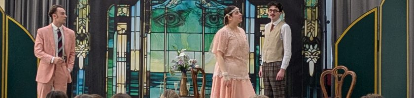 Drei Schauspieler stehen auf einer Bühne, während das Publikum im Vordergrund sitzt. Die beiden männlichen Darsteller tragen Anzüge in Pastellfarben, während die weibliche Darstellerin ein elegantes, helles Kleid trägt. Im Hintergrund ist ein dekoratives, farbiges Fenster zu sehen, das die Bühne umrahmt.