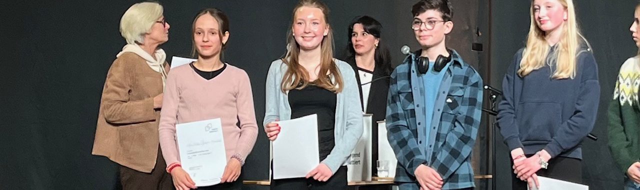 Gruppenszene mit mehreren Personen auf einer Bühne, die Urkunden oder Preise halten. Im Hintergrund sind weitere Personen zu sehen. Die Atmosphäre wirkt feierlich und stolz.