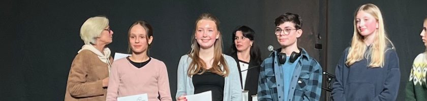 Gruppenszene mit mehreren Personen auf einer Bühne, die Urkunden oder Preise halten. Im Hintergrund sind weitere Personen zu sehen. Die Atmosphäre wirkt feierlich und stolz.