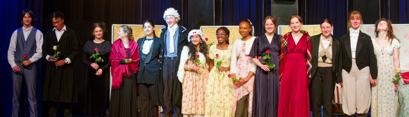 Eine Gruppe von zwölf Personen steht auf einer Bühne. Sie tragen historische Kostüme und halten jeweils eine Blume in der Hand. Im Hintergrund sind zwei große Lautsprecher und eine dunkle Vorhangwand zu sehen. Die Personen lächeln und scheinen am Ende einer Aufführung zu stehen.