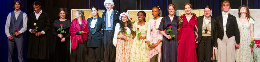 Eine Gruppe von zwölf Personen steht auf einer Bühne. Sie tragen historische Kostüme und halten jeweils eine Blume in der Hand. Im Hintergrund sind zwei große Lautsprecher und eine dunkle Vorhangwand zu sehen. Die Personen lächeln und scheinen am Ende einer Aufführung zu stehen.