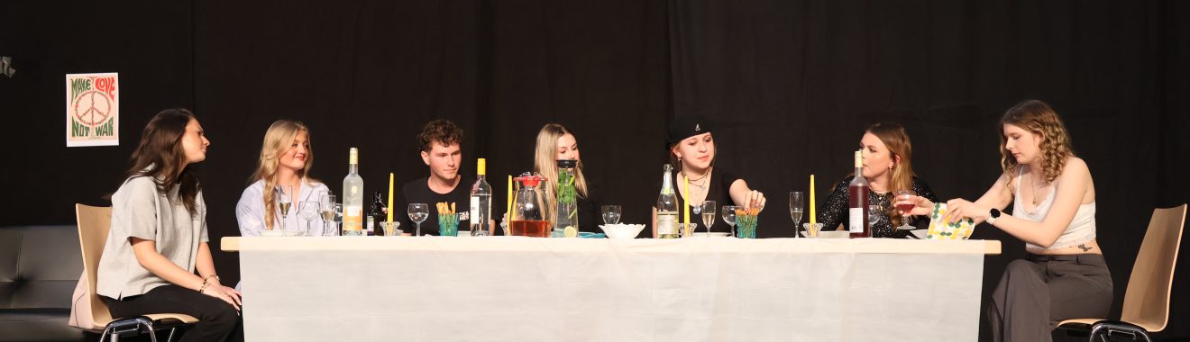 Acht Personen sitzen an einem langen Tisch, der mit verschiedenen Getränken und Speisen gedeckt ist. Die Szene wirkt gesellig, mit einer schwarzen Hintergrundwand. Einige der Personen sprechen miteinander, während andere auf den Tisch schauen. Der Raum ist hell beleuchtet, und im Hintergrund sind Stühle sichtbar.