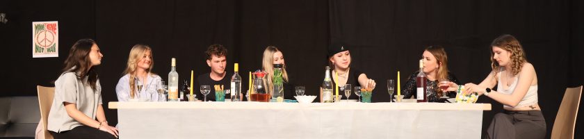 Acht Personen sitzen an einem langen Tisch, der mit verschiedenen Getränken und Speisen gedeckt ist. Die Szene wirkt gesellig, mit einer schwarzen Hintergrundwand. Einige der Personen sprechen miteinander, während andere auf den Tisch schauen. Der Raum ist hell beleuchtet, und im Hintergrund sind Stühle sichtbar.