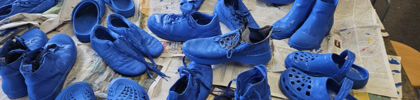 Eine Vielzahl von Schuhen in verschiedenen Formen und Stilen, die alle in leuchtendem Blau lackiert sind. Die Schuhe sind auf einer Tischoberfläche angeordnet, die mit Zeitungen bedeckt ist.