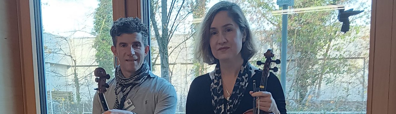 Zwei Personen sitzen auf einer Fensterbank. Der Mann links trägt ein graues Sweatshirt und schwarze Hose, während die Frau rechts ein schwarzes Kleid und schwarze Strumpfhosen trägt. Beide halten Geigen in der Hand. Im Hintergrund sind große Fenster zu sehen, durch die Tageslicht hereinfällt.