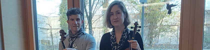 Zwei Personen sitzen auf einer Fensterbank. Der Mann links trägt ein graues Sweatshirt und schwarze Hose, während die Frau rechts ein schwarzes Kleid und schwarze Strumpfhosen trägt. Beide halten Geigen in der Hand. Im Hintergrund sind große Fenster zu sehen, durch die Tageslicht hereinfällt.