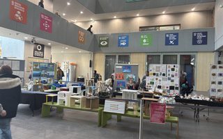 Eine Innenansicht eines Veranstaltungsraums mit mehreren Tischen, die mit Informationsmaterialien und Ausstellungsstücken bedeckt sind. Menschen bewegen sich im Raum, und an den Wänden hängen verschiedene Plakate mit Symbolen. Der Raum hat hohe Decken und große Fenster, die natürliches Licht hereinlassen.