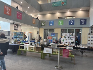 Eine Innenansicht eines Veranstaltungsraums mit mehreren Tischen, die mit Informationsmaterialien und Ausstellungsstücken bedeckt sind. Menschen bewegen sich im Raum, und an den Wänden hängen verschiedene Plakate mit Symbolen. Der Raum hat hohe Decken und große Fenster, die natürliches Licht hereinlassen.