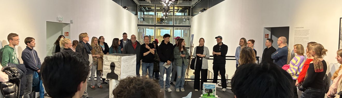 Eine Gruppe von Menschen steht in einem modernen Ausstellungsraum. Im Vordergrund sind einige Personen zu sehen, die sich um einen Tisch mit Kunstobjekten versammelt haben. Im Hintergrund stehen weitere Besucher, die auf eine Präsentation oder Ansprache warten. Die Wände sind weiß, und die Decke hat sichtbare Strukturen. Es gibt eine helle Beleuchtung und große Fenster, die Tageslicht hereinlassen.