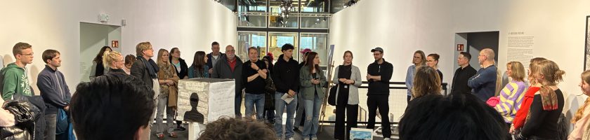 Eine Gruppe von Menschen steht in einem modernen Ausstellungsraum. Im Vordergrund sind einige Personen zu sehen, die sich um einen Tisch mit Kunstobjekten versammelt haben. Im Hintergrund stehen weitere Besucher, die auf eine Präsentation oder Ansprache warten. Die Wände sind weiß, und die Decke hat sichtbare Strukturen. Es gibt eine helle Beleuchtung und große Fenster, die Tageslicht hereinlassen.