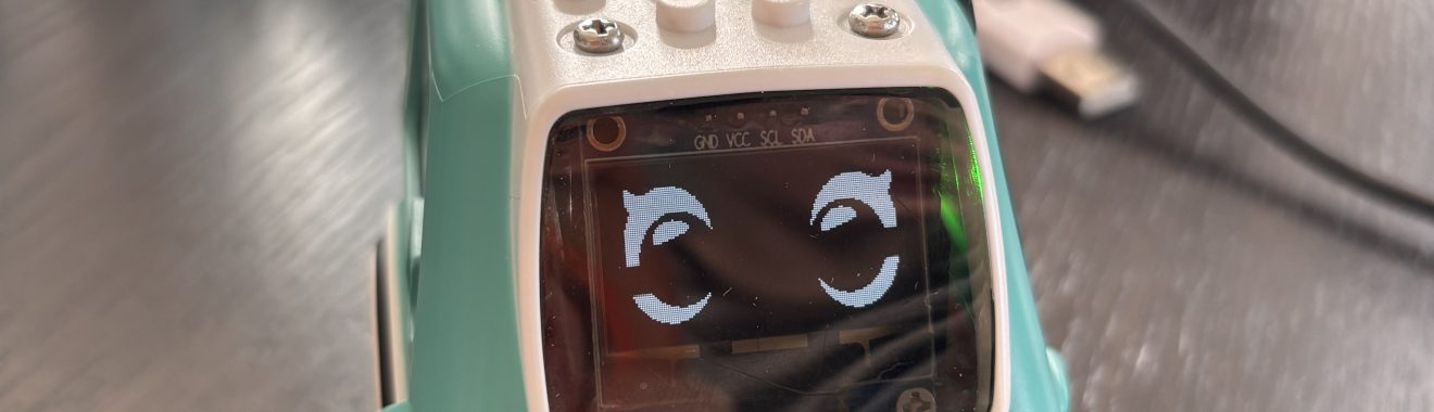 Ein kleines, mintgrünes Spielzeugauto mit einem lächelnden Gesicht auf dem Bildschirm. Das Auto hat eine weiße Unterseite und einige Knöpfe auf dem Dach. Im Hintergrund sind unscharfe Gegenstände und Kabel zu sehen.