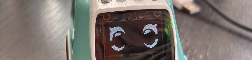 Ein kleines, mintgrünes Spielzeugauto mit einem lächelnden Gesicht auf dem Bildschirm. Das Auto hat eine weiße Unterseite und einige Knöpfe auf dem Dach. Im Hintergrund sind unscharfe Gegenstände und Kabel zu sehen.