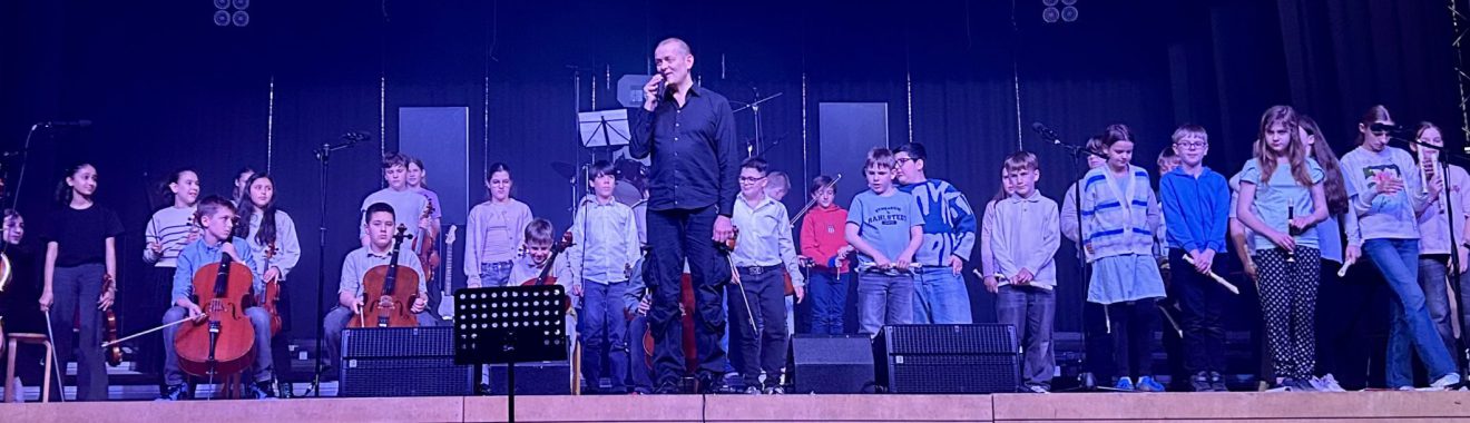 Eine Gruppe von Musikern und Schülern steht auf einer Bühne. Ein Mann steht im Vordergrund und spricht ins Mikrofon, während die anderen Musiker, darunter Streicher und Holzbläser, in der Reihe dahinter aufgestellt sind. Die Bühne ist beleuchtet und der Hintergrund ist dunkel. Alle Teilnehmer tragen ähnliche Kleidung.