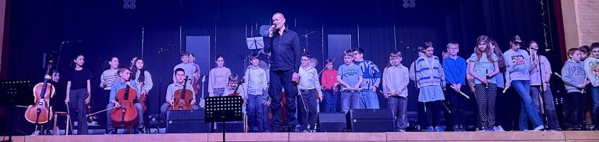 Eine Gruppe von Musikern und Schülern steht auf einer Bühne. Ein Mann steht im Vordergrund und spricht ins Mikrofon, während die anderen Musiker, darunter Streicher und Holzbläser, in der Reihe dahinter aufgestellt sind. Die Bühne ist beleuchtet und der Hintergrund ist dunkel. Alle Teilnehmer tragen ähnliche Kleidung.