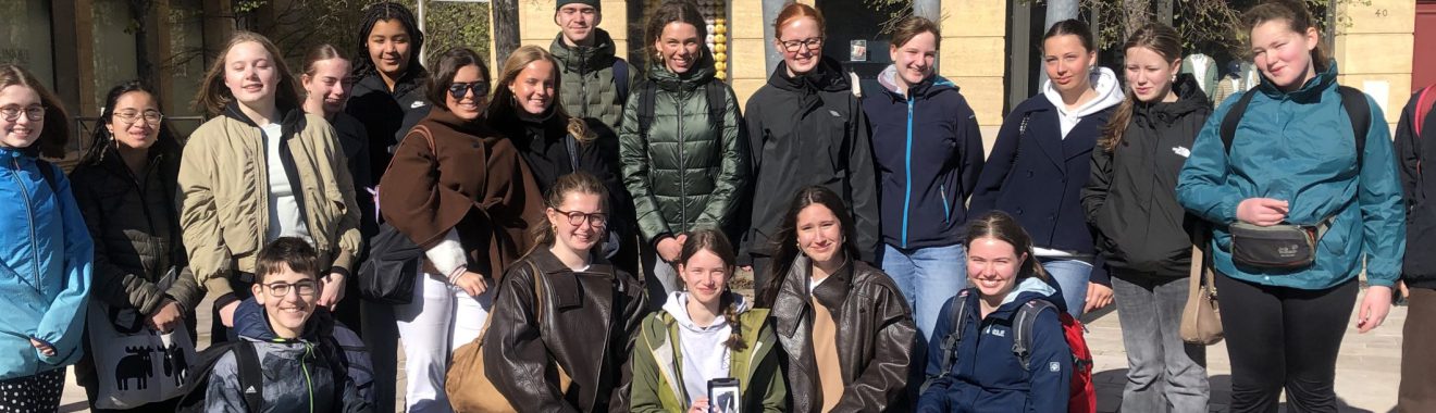 Eine Gruppe von etwa 30 Personen steht auf einem Platz vor mehreren Gebäuden. Die Teilnehmer sind in verschiedenen Winterjacken und Freizeitkleidung gekleidet. Einige Personen lächeln in die Kamera, während andere entspannt schauen. Bäume und ein blauer Himmel sind im Hintergrund sichtbar. Die Szene vermittelt eine freundliche, gemeinschaftliche Atmosphäre.