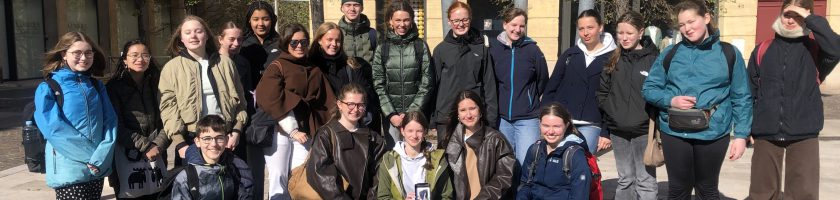 Eine Gruppe von etwa 30 Personen steht auf einem Platz vor mehreren Gebäuden. Die Teilnehmer sind in verschiedenen Winterjacken und Freizeitkleidung gekleidet. Einige Personen lächeln in die Kamera, während andere entspannt schauen. Bäume und ein blauer Himmel sind im Hintergrund sichtbar. Die Szene vermittelt eine freundliche, gemeinschaftliche Atmosphäre.