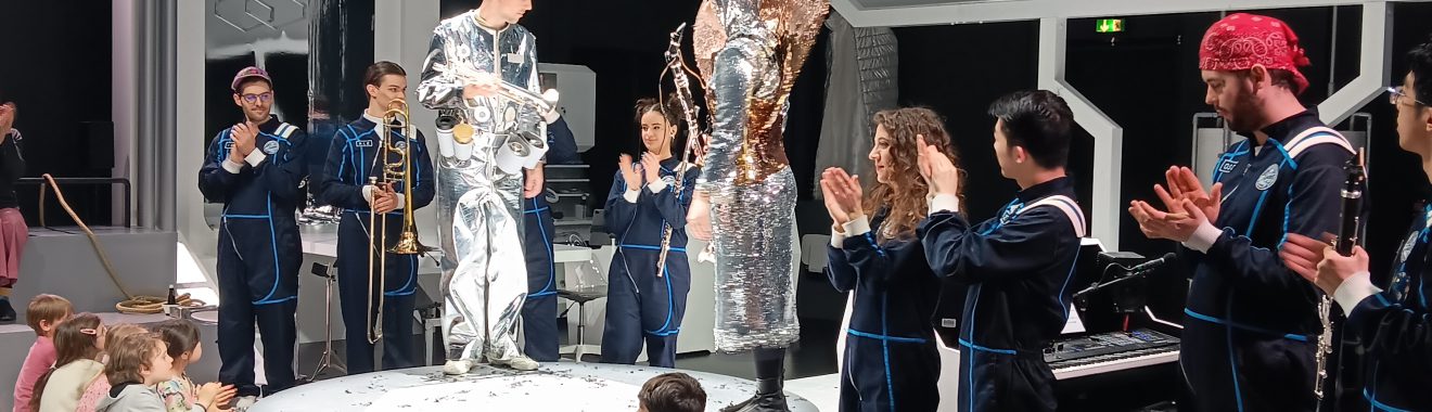 Eine Gruppe von Kindern sitzt auf einer Plattform und applaudiert. Auf der Bühne stehen mehrere Performer in auffälligen Kostümen, darunter ein silberner Anzug. Im Hintergrund sind weitere Künstler und eine Klavierbank zu sehen. Die Szene wirkt lebhaft und festlich, mit einem modernen Bühnenbild.