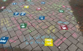 Bunte Kreidezeichnungen auf einem Pflasterboden, die hexagonale Felder in verschiedenen Farben zeigen. Jedes Feld enthält Symbole oder Wörter, die Wünsche oder Themen darstellen. Zwischen den Feldern sind weitere Kreidezeichnungen mit bunten Schriftzügen zu sehen.