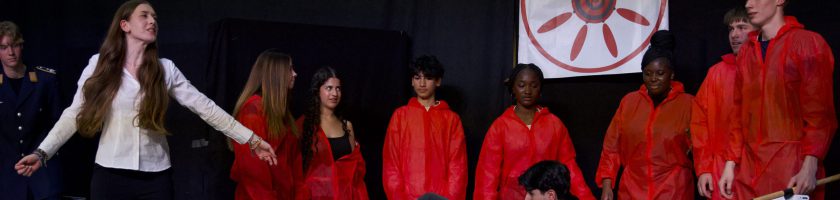 Eine Gruppe von Personen steht auf einer Bühne. Die meisten tragen rote Jacken. Eine Person mit langen Haaren in einem weißen Hemd steht im Vordergrund und gestikuliert. Im Hintergrund sind weitere Personen zu sehen, die aufmerksam schauen. Auf der Bühne ist ein großes, kreisförmiges Symbol oder Banner mit einem Sonnenmotiv angebracht. Die Atmosphäre wirkt dramatisch und theatralisch.