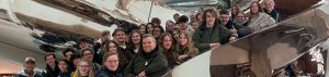 Gruppe von Menschen, die lächelnd in einem modernen, reflektierenden Raum posiert. Im Hintergrund sind gewundene, metallische Strukturen sichtbar.